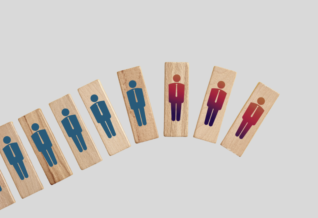 An image shows seven wooden domino-like blocks arranged in a slightly curved, falling sequence from left to right. Each block features a stencil-like illustration of a person in a suit and tie. The figures on the left four blocks are colored blue, while the figures on the right three blocks are colored in a gradient from reddish-orange to purple-red. The background is a solid light gray. The arrangement suggests a chain reaction or domino effect involving people or personnel.